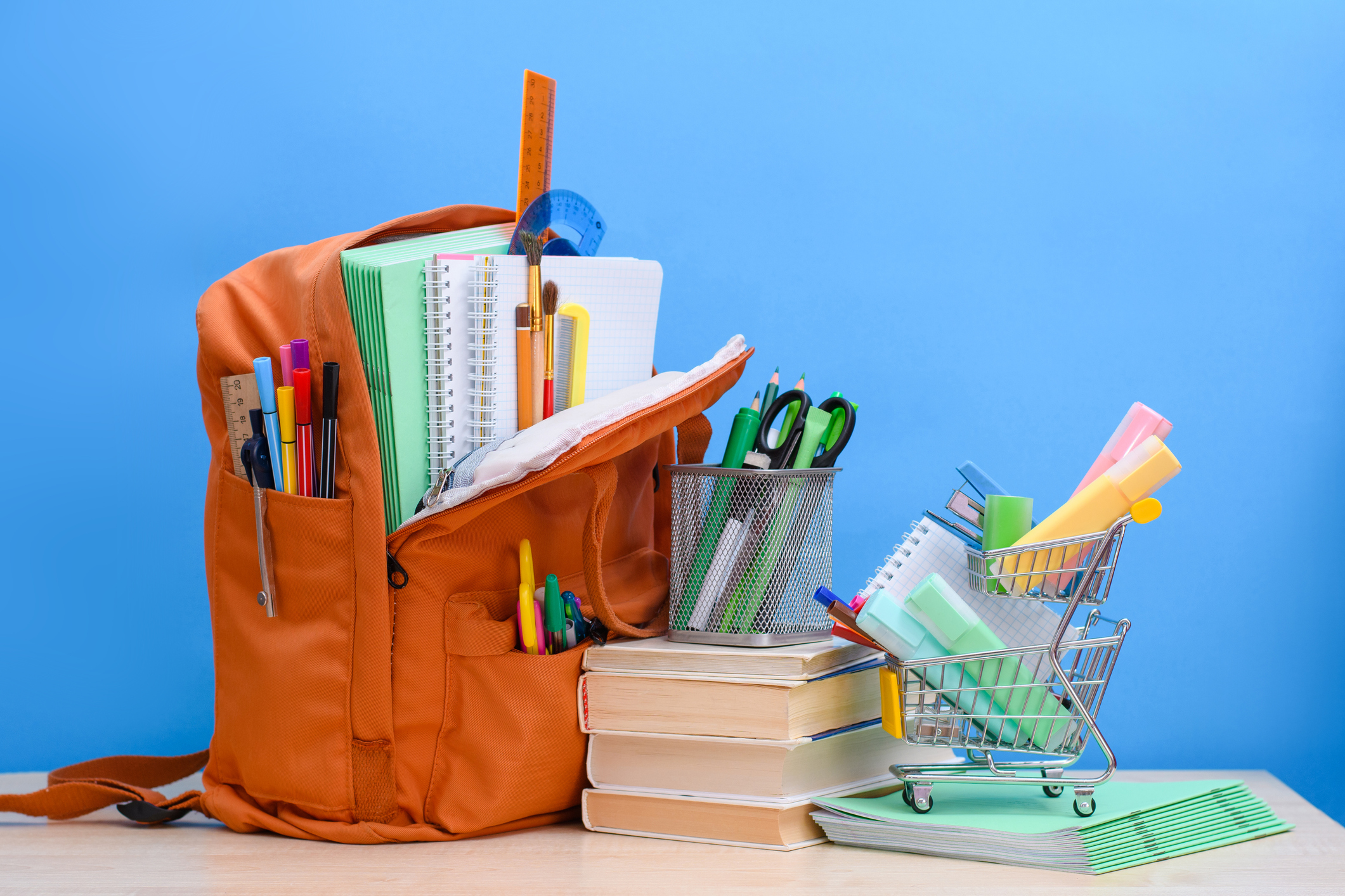 Orange school backpack full of school supplies and a supermarket basket with office supplies on a blue background.